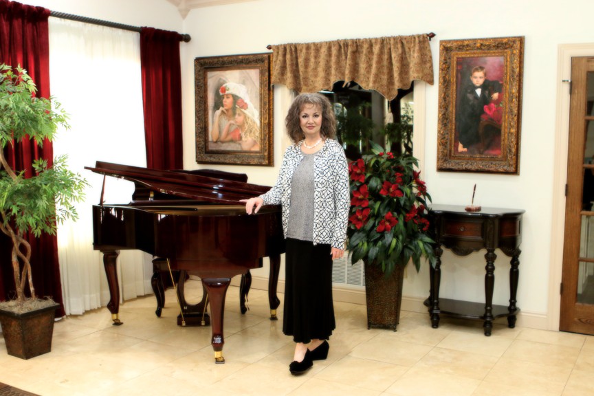 Tracy R. Lee standing beside a grand piano in a room displaying her commissioned portrait artwork.